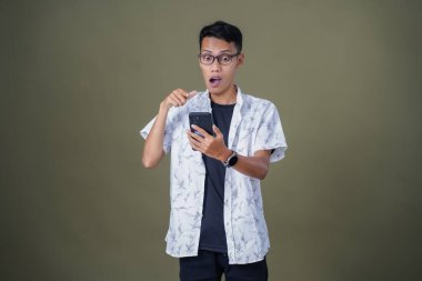 Gündelik gözlüklü genç Asyalı adam akıllı telefonu işaret ederken şaşırdı. Ordu yeşil arka planında izole edilmiş..