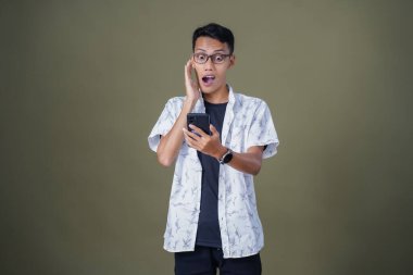 Gündelik gözlüklü genç Asyalı adam akıllı telefonu işaret ederken şaşırdı. Ordu yeşil arka planında izole edilmiş..