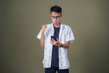 Gündelik gözlüklü genç Asyalı adam akıllı telefonu işaret ederken şaşırdı. Ordu yeşil arka planında izole edilmiş..