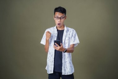 Gündelik gözlüklü genç Asyalı adam akıllı telefonu işaret ederken şaşırdı. Ordu yeşil arka planında izole edilmiş..