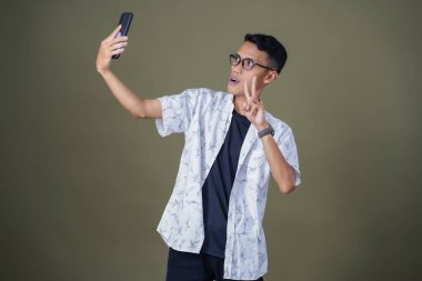 Gündelik gözlüklü genç Asyalı adam neşeli yüzüyle selfie çekiyor. Ordu yeşil arka planında izole edilmiş..