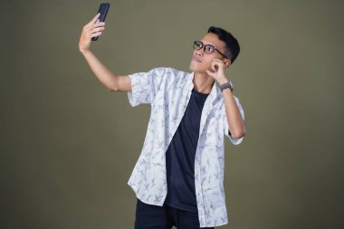 Gündelik gözlüklü genç Asyalı adam neşeli yüzüyle selfie çekiyor. Ordu yeşil arka planında izole edilmiş..