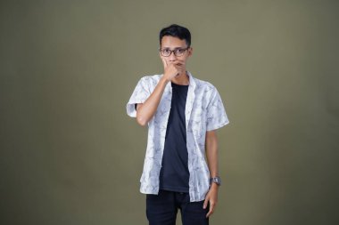 Gündelik gözlüklü genç bir Asyalı. Şaşırmış bir ifade sergiliyor. Ağzını eliyle kapatıyor. Ordu yeşil arka planında izole edilmiş..