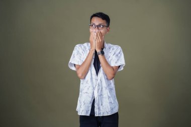 Gündelik gözlüklü genç bir Asyalı. Şaşırmış bir ifade sergiliyor. Ağzını eliyle kapatıyor. Ordu yeşil arka planında izole edilmiş..