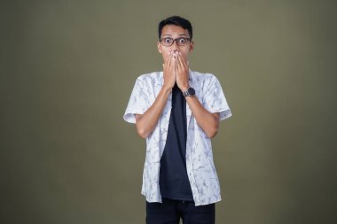 Gündelik gözlüklü genç bir Asyalı. Şaşırmış bir ifade sergiliyor. Ağzını eliyle kapatıyor. Ordu yeşil arka planında izole edilmiş..