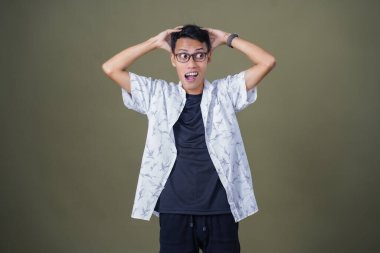 Gözlüklü genç Asyalı adam kafasını tutarken şok olmuş gibi görünüyor. Ordu yeşil arka planında izole edilmiş..