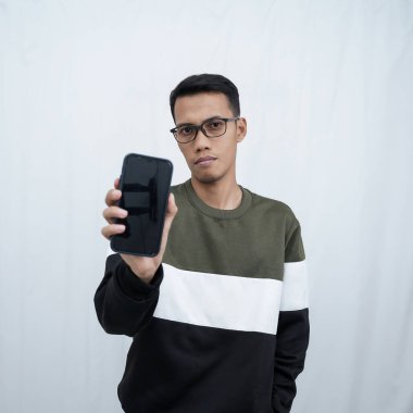 Gözlüklü, süveter giyen genç bir Asyalı akıllı telefon ekranını işaret ediyor. Beyaz bir arkaplanda izole edilmiş.