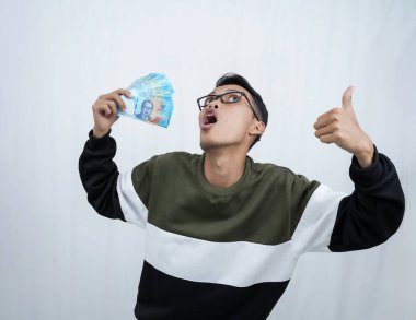 Gözlüklü Asyalı genç bir adam para taşırken mutlu bir ifadeyle kazak giyiyor, Endonezya parası. Beyaz arkaplanda izole.