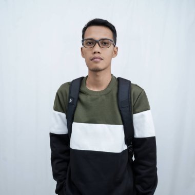 Hala okulda gözlüklü, süveter giyen ve çanta taşıyan genç bir Asyalı dik, havalı, gülümseyen bir poz veriyor. Beyaz bir arkaplanda izole edilmiş.