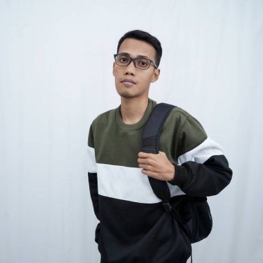 Hala okulda gözlüklü, süveter giyen ve çanta taşıyan genç bir Asyalı dik, havalı, gülümseyen bir poz veriyor. Beyaz bir arkaplanda izole edilmiş.