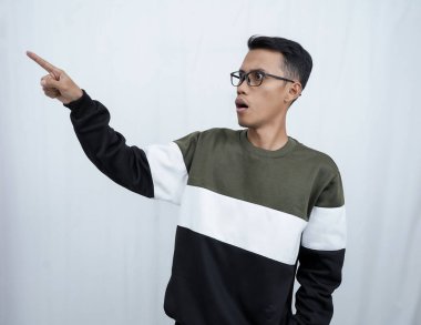 Gözlüklü genç bir Asyalı süveter giyiyor ve boş alanı gösterirken şok olmuş, şaşırmış bir ifade takınıyor. Beyaz bir arkaplanda izole edilmiş.