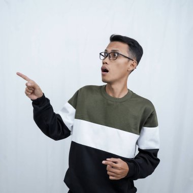 Gözlüklü genç bir Asyalı süveter giyiyor ve boş alanı gösterirken şok olmuş, şaşırmış bir ifade takınıyor. Beyaz bir arkaplanda izole edilmiş.