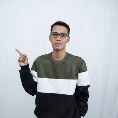 Gözlüklü, süveter giyen genç bir Asyalı boş bir yeri işaret ederken mutlu bir şekilde gülümsüyor. Beyaz bir arkaplanda izole edilmiş.