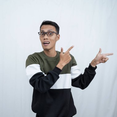 Gözlüklü genç bir Asyalı süveter giyiyor ve boş alanı gösterirken şok olmuş, şaşırmış bir ifade takınıyor. Beyaz bir arkaplanda izole edilmiş.