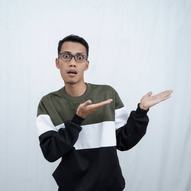 Gözlüklü genç bir Asyalı süveter giyiyor ve boş alanı gösterirken şok olmuş, şaşırmış bir ifade takınıyor. Beyaz bir arkaplanda izole edilmiş.