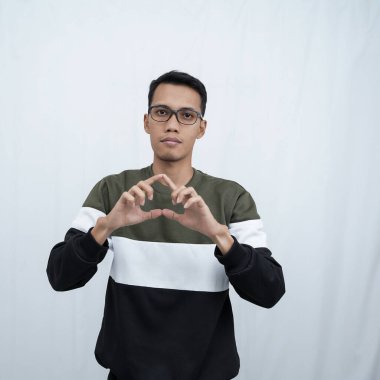 Gözlüklü, süveter giyen genç bir Asyalı elleriyle aşk işareti yaparken gülümsüyor. Beyaz arkaplanda izole.