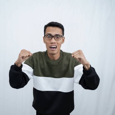 Gözlüklü, mutlu, heyecanlı ve muzaffer bir ifadeyle süveter giyen genç bir Asyalı yumruğunu kaldırıp sıkarken. Beyaz arkaplanda izole.