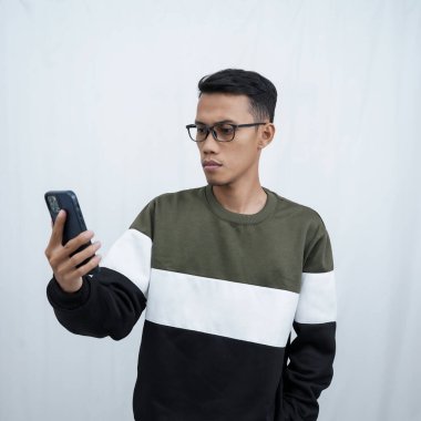 Gözlüklü, süveter giyen, mutlu bir ifadeyle akıllı telefona bakarken şaşıran genç bir Asyalı. Beyaz bir arkaplanda izole edilmiş.