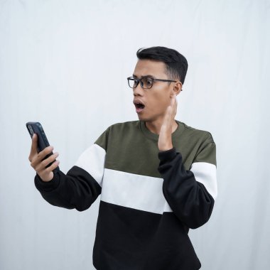 Gözlüklü, süveter giyen, mutlu bir ifadeyle akıllı telefona bakarken şaşıran genç bir Asyalı. Beyaz bir arkaplanda izole edilmiş.