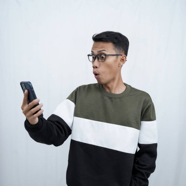 Gözlüklü, süveter giyen, mutlu bir ifadeyle akıllı telefona bakarken şaşıran genç bir Asyalı. Beyaz bir arkaplanda izole edilmiş.