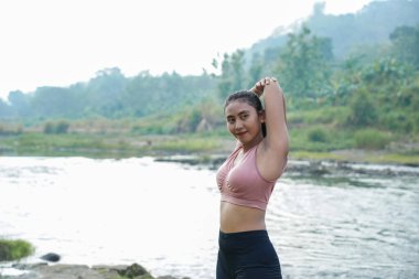 Spor kıyafetleri içinde kahverengi tenli ince bir Asyalı kız spor yapmadan önce güzel bir nehrin kenarında triseps hareketlerini geriyor..