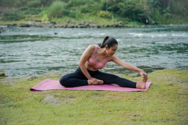 Spor kıyafetleri içinde kahverengi tenli ince bir Asyalı kız spor yapmadan önce güzel bir nehrin kenarındaki açık havada dizlerini esnetiyor..