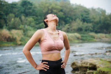 Sıska, bronz tenli bir Asyalı kız spor kıyafetleri içinde güzel bir nehrin kenarında boyun bükme egzersizleri yapıyor..