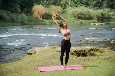 Zihinsel ve fiziksel sağlık. Sıska, bronz tenli Asyalı bir kız sabah sakin bir nehirde yoga yapıyor. Kadın Tadasana pozunda elleri başının üstünde duruyor..