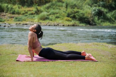 Zihinsel ve fiziksel sağlık. İnce, bronz tenli, spor giyimli Asyalı bir kadın sabahları güzel bir nehir kıyısında yoga yapıyor. Kadın kobra pozu veriyor..