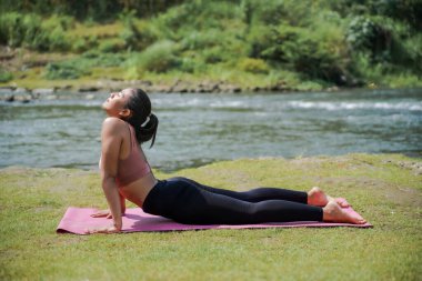 Zihinsel ve fiziksel sağlık. İnce, bronz tenli, spor giyimli Asyalı bir kadın sabahları güzel bir nehir kıyısında yoga yapıyor. Kadın kobra pozu veriyor..