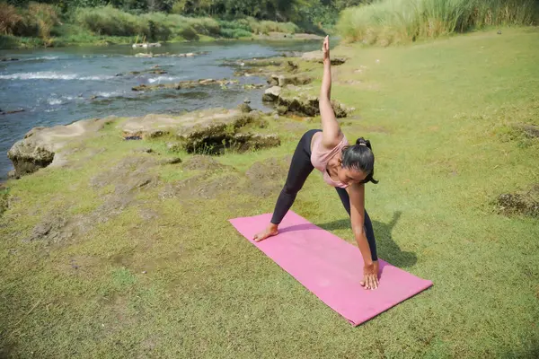 Zihinsel ve fiziksel sağlık. Sıska, bronz tenli Asyalı bir kız sabah sakin bir nehirde yoga yapıyor. Kadın Genişletilmiş Üçgen 'de duruyor Kolları aşağıda ve yukarıda poz veriyor.
