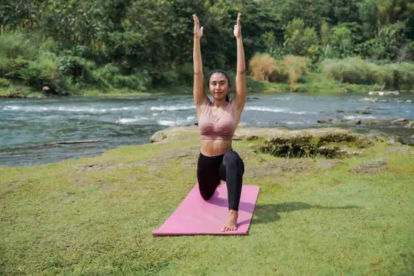 Zihinsel ve fiziksel sağlık. İnce, bronz tenli, atletik kıyafetli Asyalı bir kadın sabahları güzel bir nehir kıyısında yoga yapıyor. Kadın savaşçı pozu veriyor, ya da Virabhadrasana.