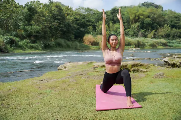 Zihinsel ve fiziksel sağlık. İnce, bronz tenli, atletik kıyafetli Asyalı bir kadın sabahları güzel bir nehir kıyısında yoga yapıyor. Kadın savaşçı pozu veriyor, ya da Virabhadrasana.