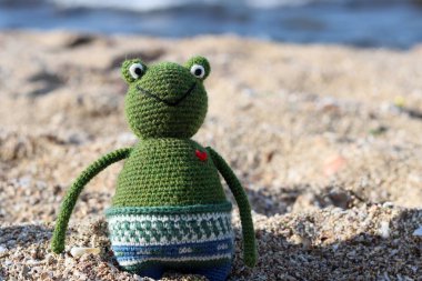 Crochet Frog toy on the beach. Cute amigurumi toy sitting on sand, blue sea on a background. 