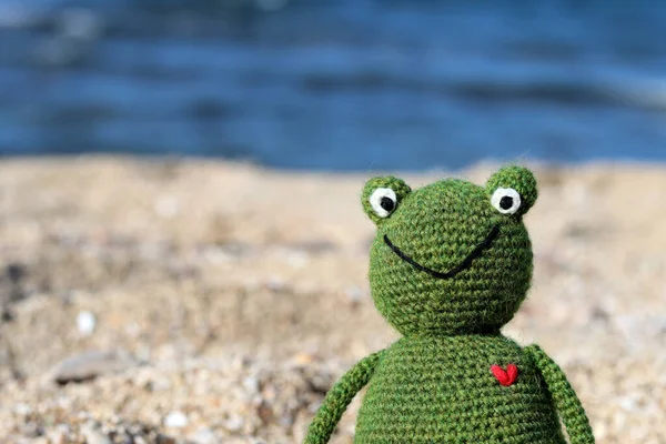 Crochet Frog toy on the beach. Cute amigurumi toy sitting on sand, blue sea on a background.