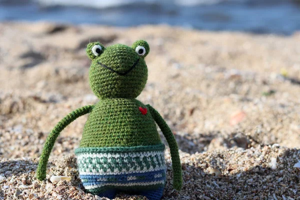 Crochet Frog toy on the beach. Cute amigurumi toy sitting on sand, blue sea on a background.