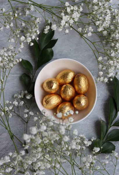 Chocolate Easter Egg Wrapped in Gold Foil. White spring flowers and Ester sweets top view photo. Bright spring still life.