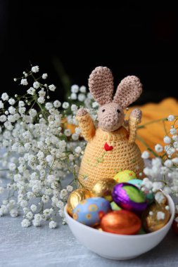 Tığ işi tavşan ve masada bir kase Paskalya tatlısı. Çikolatalı yumurtalar, çiçekler ve oyuncaklarla dolu renkli bir hayat. Paskalya kutlaması kavramı. 