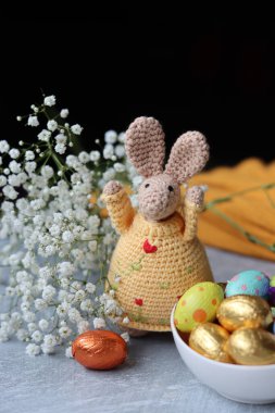 Tığ işi tavşan ve masada bir kase Paskalya tatlısı. Çikolatalı yumurtalar, çiçekler ve oyuncaklarla dolu renkli bir hayat. Paskalya kutlaması kavramı. 