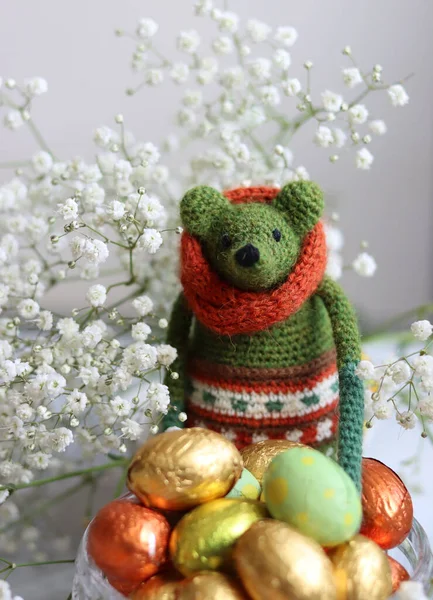 Crochet green toy, chocolate eggs and white flowers on a table. Handmade gift for Easter. Colorful still life photo with oil wrapped sweets. Easter celebration.