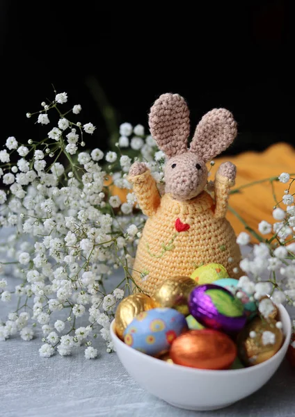 Crochet bunny and bowl of Easter sweets on a table. Colorful still life with chocolate eggs, flowers and toy. Easter celebration concept.