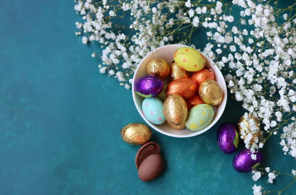Colorful still life with chocolate eggs and gypsophila flowers. Easter greeting card. Small wrapped chocolate eggs on a decorative plate. Easter sweets close up photo with copy space.