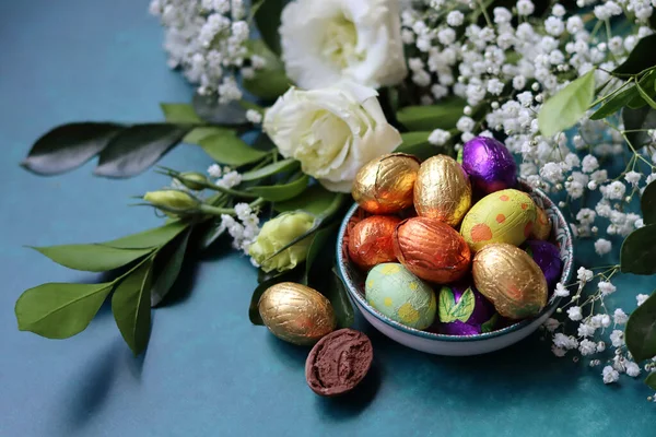 Colorful still life with chocolate eggs and gypsophila flowers. Easter greeting card. Small wrapped chocolate eggs on a decorative plate. Easter sweets close up photo with copy space.