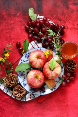 Kırmızı arka planda kırmızı elma ve üzüm ve bir fincan bal. Yahudi yeni yılı Rosh Hashana kutlaması. Mevsimlik meyve ve bal ile sakin bir hayat. 