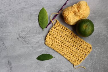 Tığ işi eşarp işleniyor. Örgü kancası, sarı pamuk ipliği, kireç ve yeşil yapraklar gri arka planda, üst manzara 