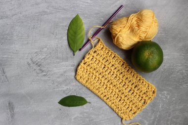 Tığ işi eşarp işleniyor. Örgü kancası, sarı pamuk ipliği, kireç ve yeşil yapraklar gri arka planda, üst manzara 