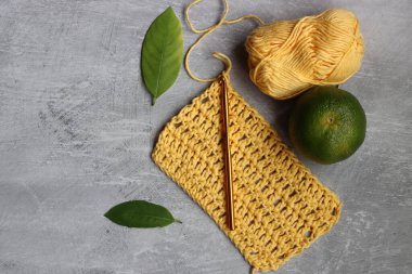 Tığ işi eşarp işleniyor. Örgü kancası, sarı pamuk ipliği, kireç ve yeşil yapraklar gri arka planda, üst manzara 