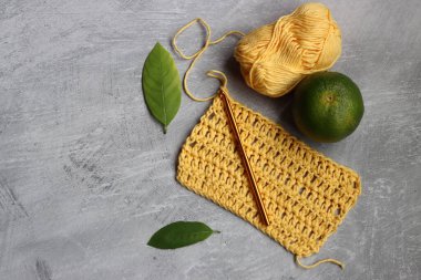 Tığ işi eşarp işleniyor. Örgü kancası, sarı pamuk ipliği, kireç ve yeşil yapraklar gri arka planda, üst manzara 