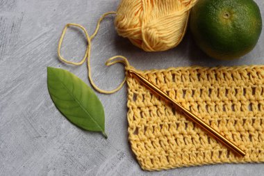 Tığ işi eşarp işleniyor. Örgü kancası, sarı pamuk ipliği, kireç ve yeşil yapraklar gri arka planda, üst manzara 