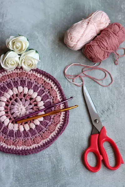 Crochet hook with cotton thread, scissors and roses on grey table, closeup photo of handmade pattern. Leisure and hobby concept. Space for text.